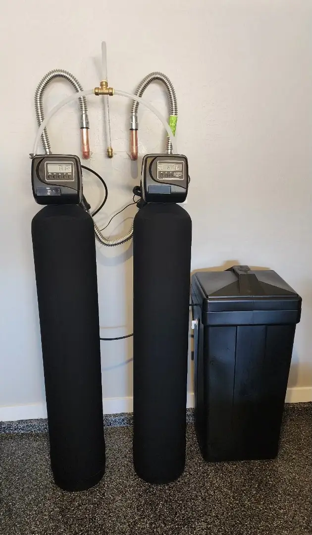 Two black water treatment tanks with digital control heads, copper stubs and a brine tank against a beige wall